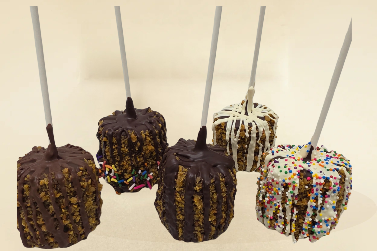 Five chocolate-dipped toffee marshmallows on sticks with various toppings against a beige background