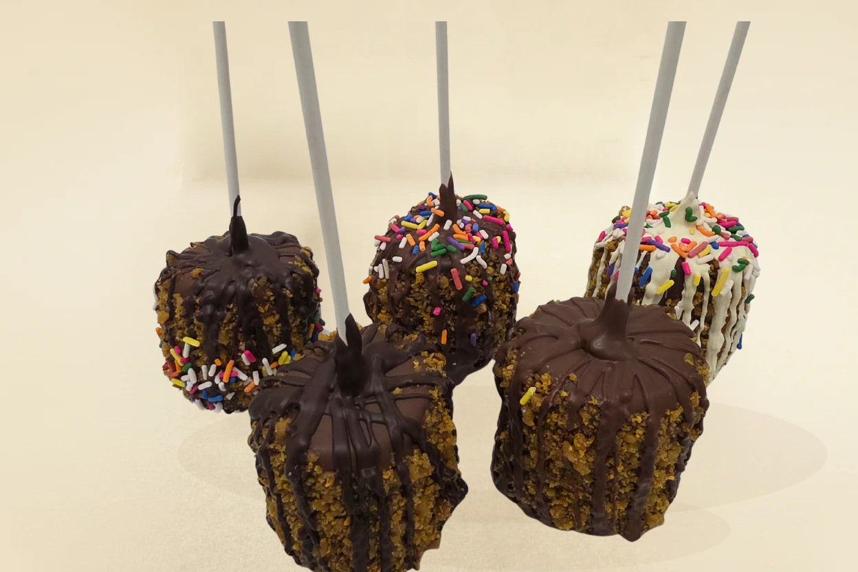 Five chocolate-dipped apples on sticks with sprinkles against a beige background